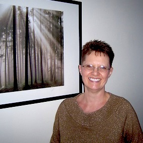 Picture of Anne Reith next to framed photograph of sunlight coming through tall trees